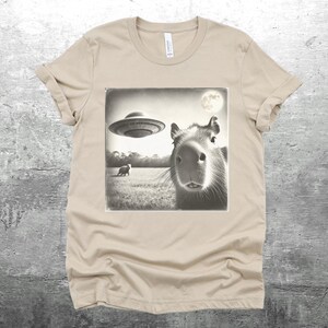 K&ouml;nnte beinhalten: Ein beiges T-Shirt mit einem Schwarzwei&szlig;bild eines Kapibaras, das in die Kamera schaut. Ein UFO fliegt &uuml;ber dem Kapibara im Bild. Das Bild hat einen Vintage-Look.