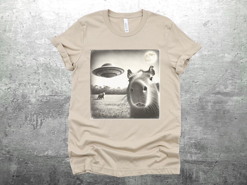 K&ouml;nnte beinhalten: Ein beiges T-Shirt mit einem Schwarzwei&szlig;bild eines Kapibaras, das in die Kamera schaut. Ein UFO fliegt &uuml;ber dem Kapibara im Bild. Das Bild hat einen Vintage-Look.