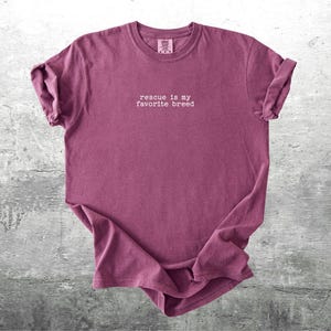 May include: A maroon t-shirt with the phrase "rescue is my favorite breed" in white, centered on the front. The shirt has rolled-up sleeves and is displayed on a gray, textured surface.
