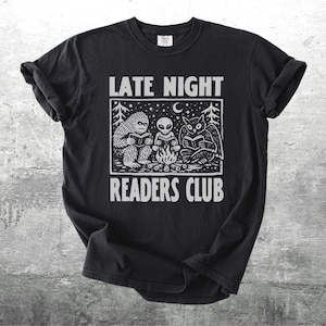 May include: Black t-shirt with a white graphic of a Bigfoot, an alien, and an owl reading around a campfire. The text "LATE NIGHT READERS CLUB" is above and below the graphic. The shirt is on a gray textured background.