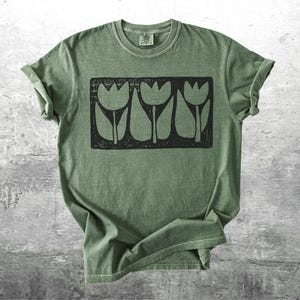 May include: Olive green t-shirt featuring a black rectangular graphic of three stylized tulip flowers. The shirt has a crew neck and short sleeves, with the sleeves rolled up. The fabric has a slightly faded, vintage look.