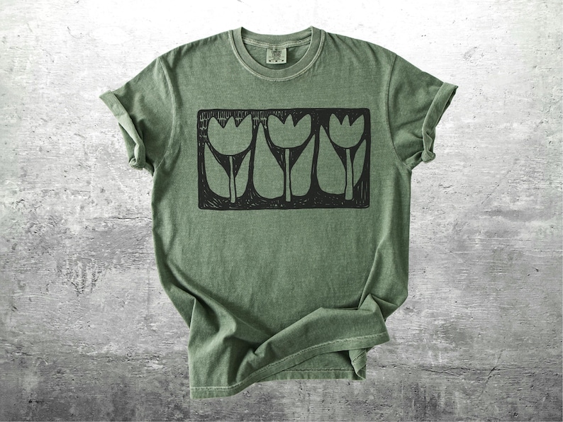 May include: Olive green t-shirt featuring a black rectangular graphic of three stylized tulip flowers. The shirt has a crew neck and short sleeves, with the sleeves rolled up. The fabric has a slightly faded, vintage look.