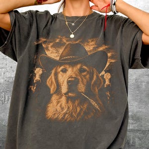 May include: Dark gray t-shirt featuring a golden retriever wearing a cowboy hat and holding a piece of wheat in its mouth. The design includes cows and a cloudy sky. The shirt has a vintage, distressed look.