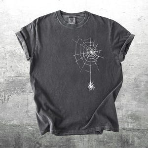 May include: Dark gray t-shirt featuring a white spider web design with a spider hanging below. The shirt has rolled-up sleeves and a crew neck. The fabric appears to be pre-washed for a vintage look. The shirt is on a gray concrete background.