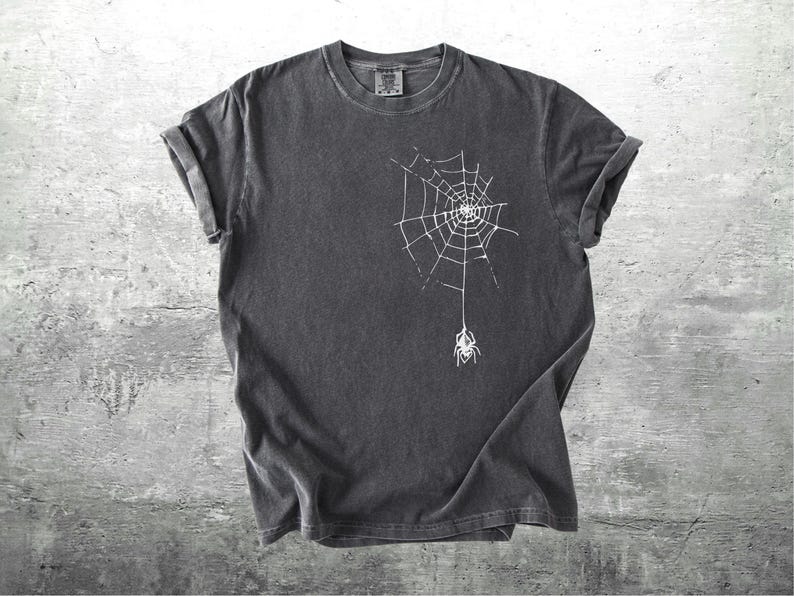 May include: Dark gray t-shirt featuring a white spider web design with a spider hanging below. The shirt has rolled-up sleeves and a crew neck. The fabric appears to be pre-washed for a vintage look. The shirt is on a gray concrete background.
