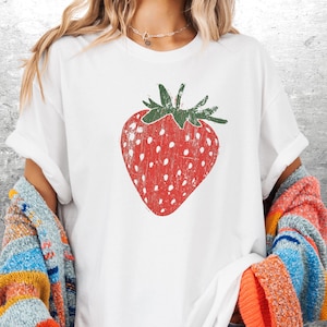 May include: White t-shirt featuring a distressed red strawberry graphic with white seeds and a green stem. The shirt is part of a clothing collection and is suitable for casual wear.