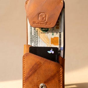 May include: A tan leather wallet with a snap closure, containing a stack of US hundred-dollar bills and a black credit card. The wallet features the text "Made with Soul" and a logo.