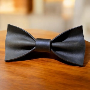 May include: A black leather bow tie with a classic butterfly shape. The bow tie is made of a single piece of leather and has a smooth, matte finish. The bow tie is sitting on a wooden surface.