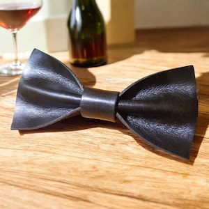 Può includere: Un papillon in pelle nera dalla forma classica a farfalla. Il papillon è su una superficie di legno, con un bicchiere e una bottiglia di vino sullo sfondo. Un accessorio formale.