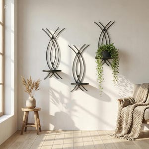 May include: Three black metal wall shelves with a unique, curved design. One shelf holds a potted plant with trailing green foliage. The shelves are mounted on a white wall, creating a modern, minimalist aesthetic.