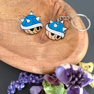 Könnte beinhalten: Zwei blaue Schlüsselanhänger in Muschelform mit weißen Stacheln und einem Cartoon-Gesicht, die auf einer Holzschale liegen. Die Schlüsselanhänger sind silberfarben und die Schale ist von getrockneten lila und grünen Blumen umgeben.