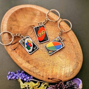 Può includere: Tre portachiavi con disegni di carte UNO. Ogni portachiavi ha una catena e un anello color argento. Le carte UNO hanno uno sfondo nero con grafica colorata e la scritta "UNO". I portachiavi sono esposti su una ciotola di legno.