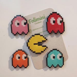 Può includere: Una collezione di personaggi pixelati di Pac-Man, tra cui fantasmi rosa, rossi, arancioni e turchese, insieme a una figura gialla di Pac-Man. I personaggi sono disposti su una scheda marrone chiaro con il testo "Folimini Créations".