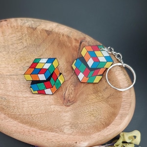 Op de afbeelding: Twee kleurrijke accessoires met een Rubik's Cube-thema. Eén is een sleutelhanger met een zilveren ring, de andere is een bijpassende speld. Beide hebben het klassieke kubusontwerp met levendige rode, blauwe, gele, groene, oranje en witte vierkanten. Ze liggen op een houten dienblad.