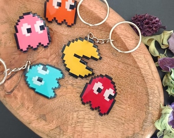May include: A collection of five pixelated keychains featuring a yellow circular character with a wedge-shaped mouth and four ghost-shaped characters in orange, pink, light blue, and red, each attached to a silver metal split ring keychain.