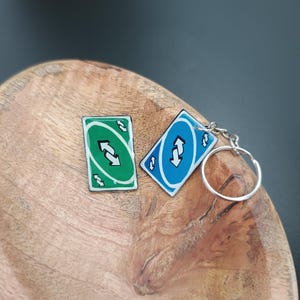 Könnte beinhalten: Zwei Schlüsselanhänger im Uno-Kartendesign. Einer ist grün mit weißen Pfeilen, der andere blau mit weißen Pfeilen und einem silbernen Schlüsselring. Die Schlüsselanhänger liegen auf einer Holzschale.