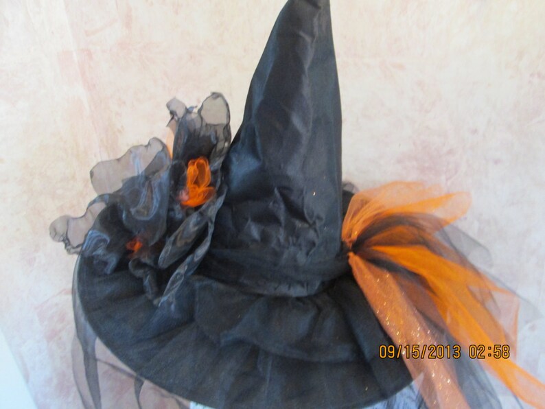 May include: Black witch hat with orange and black tulle accents. The hat has a tall pointed crown and a wide brim. The tulle is gathered into a large bow on the top of the hat.