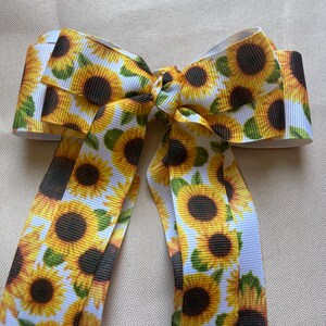 May include: A large hair bow featuring a repeating pattern of sunflowers in yellow, brown, and green on a white background. The bow is tied in the center with long, flowing tails.
