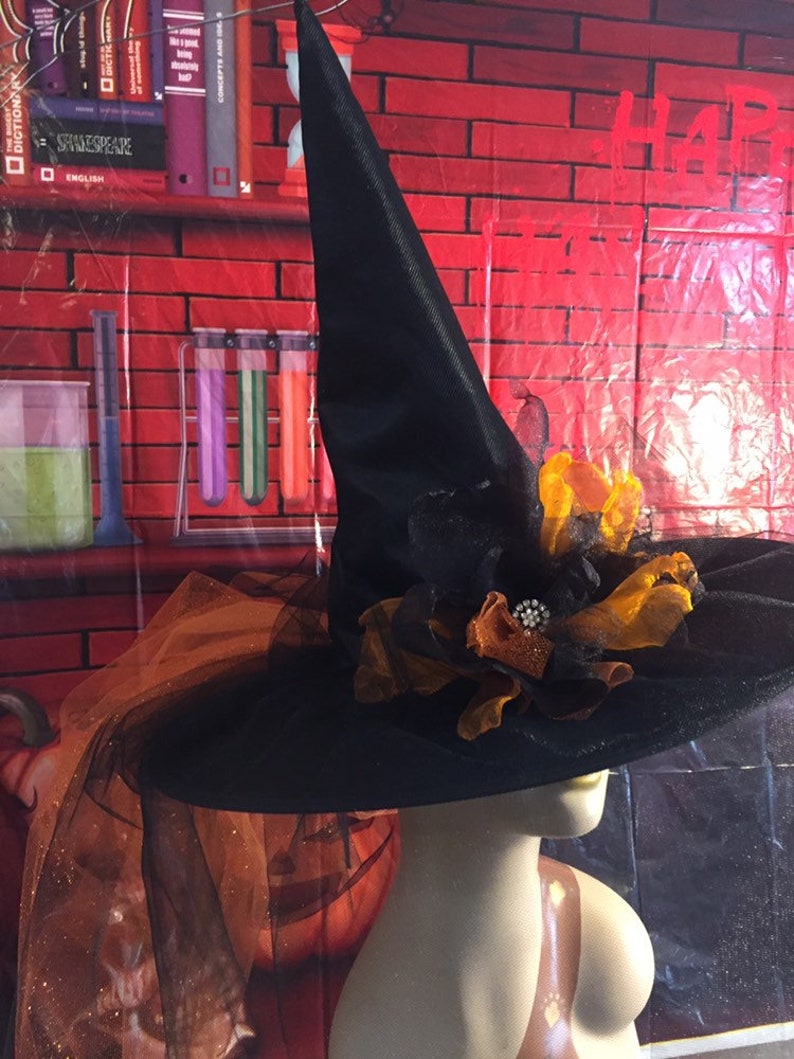 May include: A black witch hat with a wide brim and a tall pointed crown. The hat is decorated with a large orange and black flower with a rhinestone embellishment. The hat is on a mannequin against a red brick background.