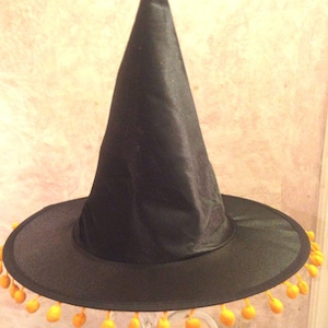 May include: A black witch hat with a tall pointed crown and a wide brim. The brim is decorated with a row of orange pom-poms.