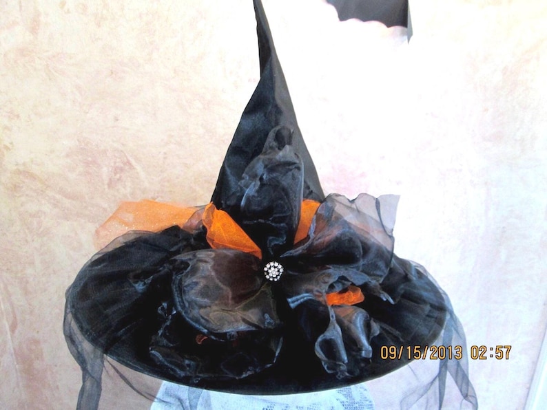 May include: A black witch hat with orange and black tulle. The hat has a large, black brim and a tall, pointed crown. A single rhinestone embellishment is centered on the front of the hat.
