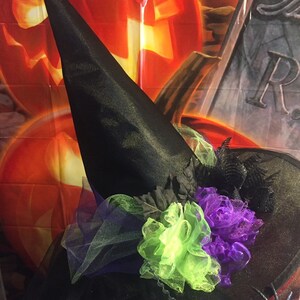 May include: A black witch's hat with a green and purple tulle flower embellishment. The hat is in front of a Halloween backdrop with pumpkins and a tombstone.