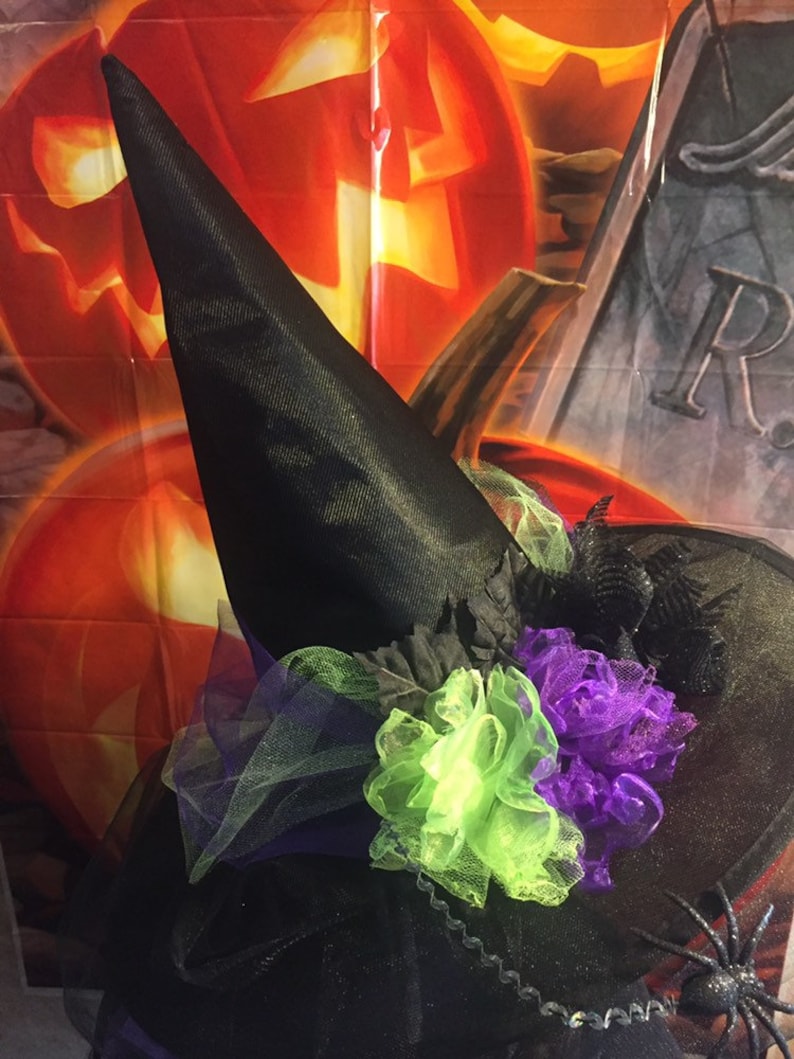 May include: A black witch's hat with a green and purple tulle flower embellishment. The hat is in front of a Halloween backdrop with pumpkins and a tombstone.