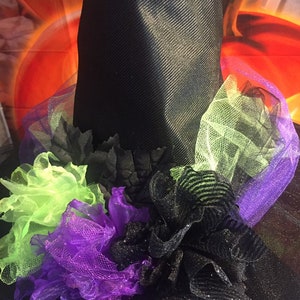 May include: A black witch hat with a wide brim and a black tulle base. The hat is decorated with green, purple, and black tulle ruffles and flowers.
