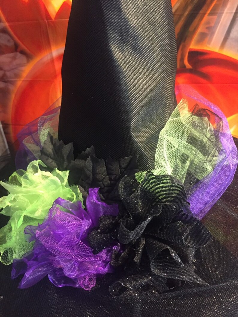 May include: A black witch hat with a wide brim and a black tulle base. The hat is decorated with green, purple, and black tulle ruffles and flowers.