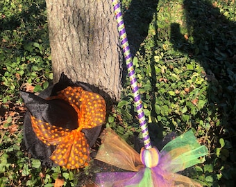 May include: A witch's broom with a purple and green striped handle and a green, purple, and orange tulle bow. The broom is leaning against a tree trunk. A black witch's hat with an orange polka dot bow is lying on the ground.