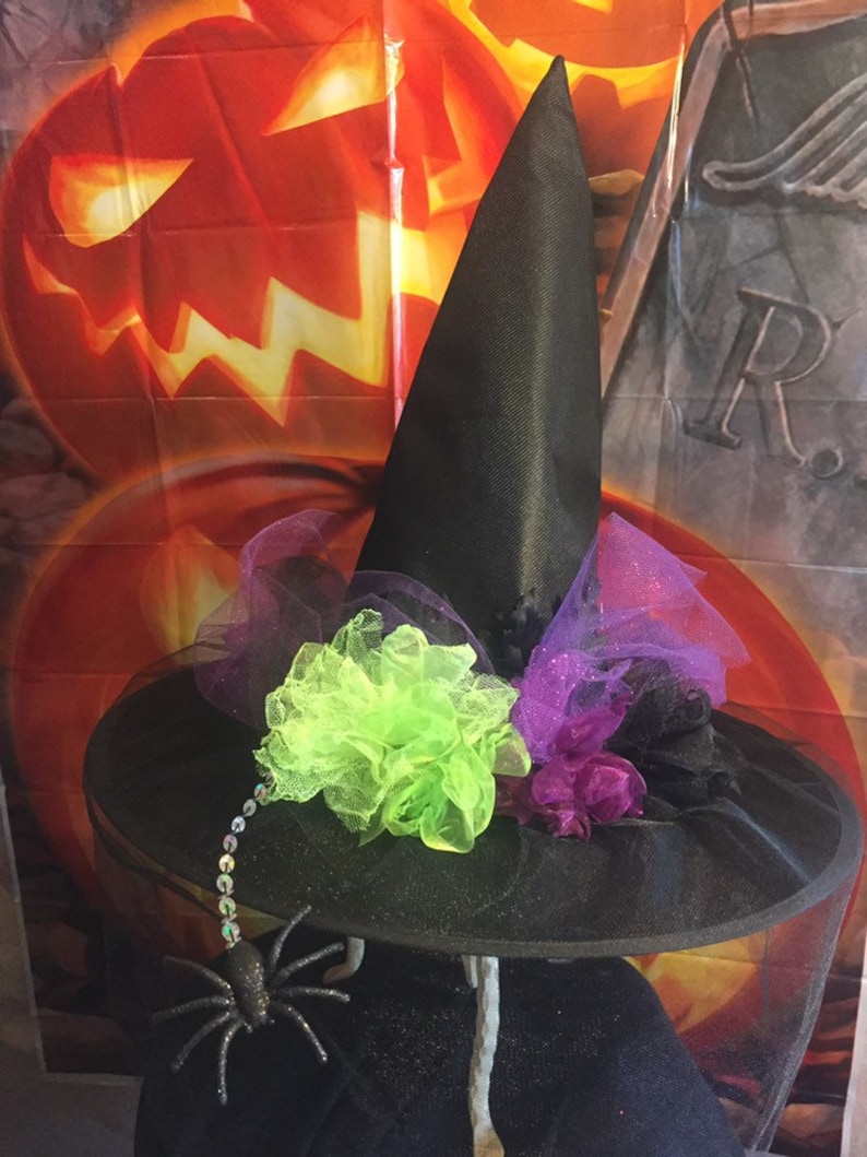 May include: Black witch hat with a wide brim, decorated with purple and green tulle, a green flower, and a black spider with silver beads.