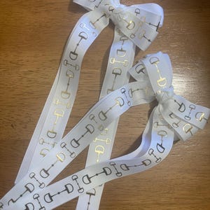 May include: White ribbon with a gold equestrian bit pattern. The ribbon is tied into bows with long, flowing tails. The gold bits are evenly spaced along the ribbon's length. The bows are arranged on a wooden surface.