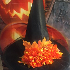 May include: A black witch hat with a wide brim and a tall pointed crown. The hat is decorated with orange and brown fall leaves and a large orange flower.