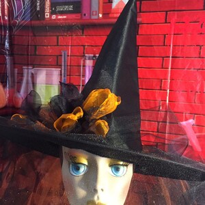 May include: A black witch's hat with a wide brim and a black veil. The hat is decorated with orange and black ribbon and a black feather. The hat is sitting on a mannequin head with a painted face.
