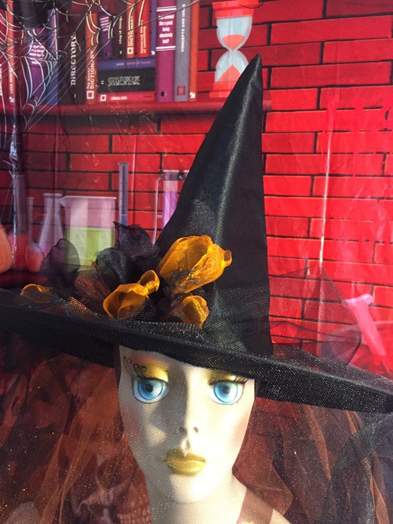 May include: A black witch's hat with a wide brim and a black veil. The hat is decorated with orange and black ribbon and a black feather. The hat is sitting on a mannequin head with a painted face.