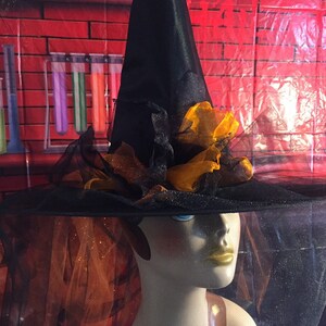 May include: A black witch's hat with a wide brim and a tall pointed crown. The hat is decorated with orange and black fabric flowers. The hat is sitting on a mannequin's head.