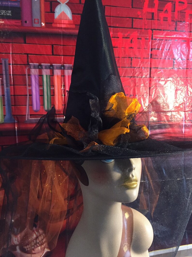 May include: A black witch's hat with a wide brim and a tall pointed crown. The hat is decorated with orange and black fabric flowers. The hat is sitting on a mannequin's head.