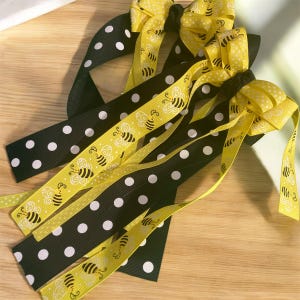May include: Several hair bows with a bee theme. The bows feature black ribbon with white polka dots and yellow ribbon with bee graphics. The bows are tied with yellow ribbon and are arranged on a wooden surface.