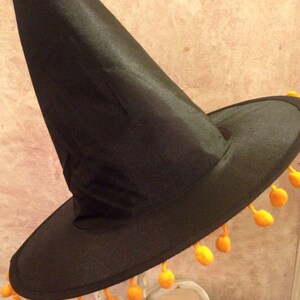 May include: A black witch hat with a wide brim and a tall pointed crown. The brim is trimmed with a row of orange pom-poms.