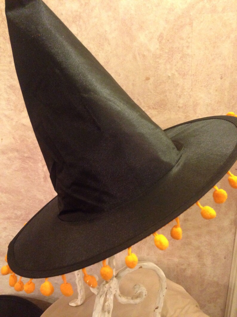 May include: A black witch hat with a wide brim and a tall pointed crown. The brim is trimmed with a row of orange pom-poms.