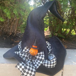 May include: A black pointed hat featuring a large black and white checkered bow at the base. Centered on the bow is a small orange jack-o'-lantern figure wearing a tiny black hat with an orange band. The hat has a soft, textured finish.