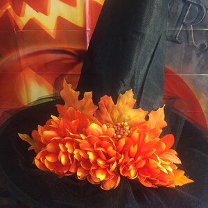 May include: A black witch hat with a wide brim and a tall pointed crown. The hat is decorated with orange fabric flowers and fall leaves.