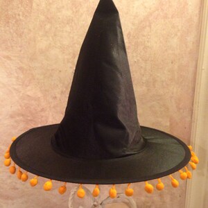 May include: A black witch hat with a wide brim and orange pom-poms around the edge. The hat is sitting on a white stand.