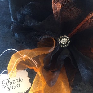 May include: A black witch hat with orange and black tulle and a rhinestone embellishment. A white tag with the text "Thank You" is attached to the hat.