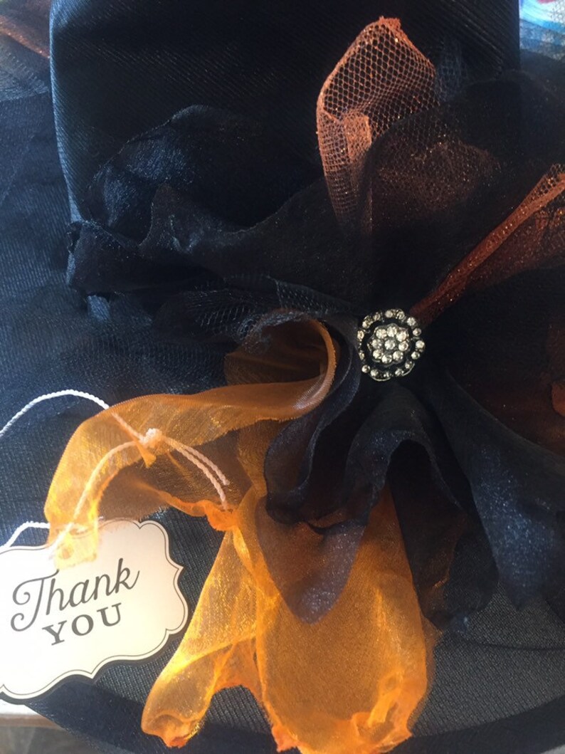 May include: A black witch hat with orange and black tulle and a rhinestone embellishment. A white tag with the text "Thank You" is attached to the hat.