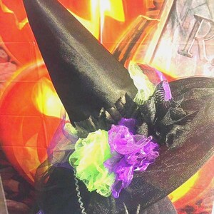 May include: A black witch's hat with a wide brim and a tall pointed crown. The hat is decorated with black tulle and green and purple flowers.