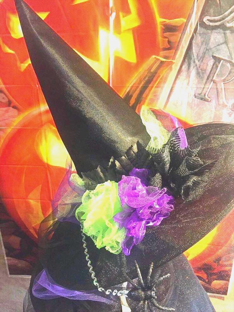 May include: A black witch's hat with a wide brim and a tall pointed crown. The hat is decorated with black tulle and green and purple flowers.