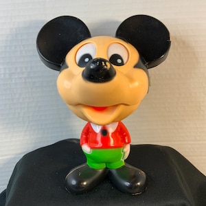 May include: A Mickey Mouse figurine with a black head and ears, a tan face, and a red shirt with green pants. The figurine has large black shoes and a smiling expression.