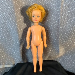 Könnte beinhalten: Eine Vintage-Puppe mit pfirsichfarbenem Körper, blondem Haar und blauen Augen. Die Puppe hat bewegliche Arme und Beine und steht auf einer schwarzen Oberfläche vor einem gemusterten Hintergrund. Zu den Gesichtszügen der Puppe gehören gemalte Augenbrauen, Wimpern und ein roter Mund.