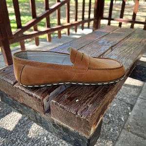 K&ouml;nnte beinhalten: Ein einzelner brauner Leder-Loafer mit Penny-Riemen-Detail, der auf einer verwitterten Holzbank liegt. Der Schuh hat eine wei&szlig; gen&auml;hte Sohle und ein klassisches Design. Der Hintergrund zeigt eine Holzkonstruktion und etwas Gr&uuml;n. Der Schuh ist etwa 25 cm lang.